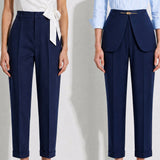 A model showcasing elegant navy blue high-waisted work trousers, featuring a unique curved overlay panel design at the waist, a slim golden metal bar buckle, pressed front creases, and chic cuffed hems, paired with a light blue button-up shirt.