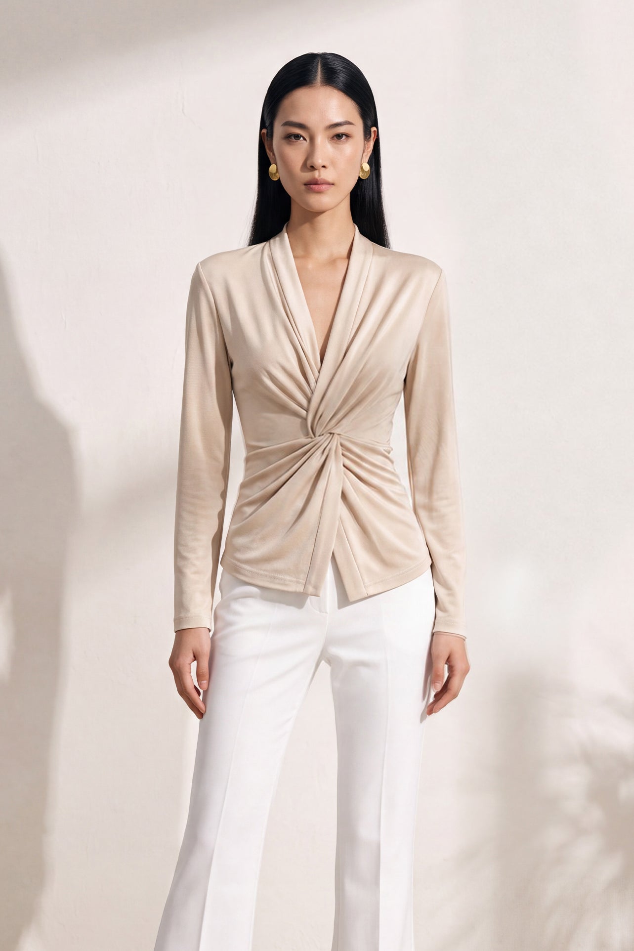 A chic Asian woman modeling an elegant champagne gold long-sleeve top featuring a sophisticated v-neckline and a stylized twist-front knot waist, beautifully paired with high-waisted white trousers.