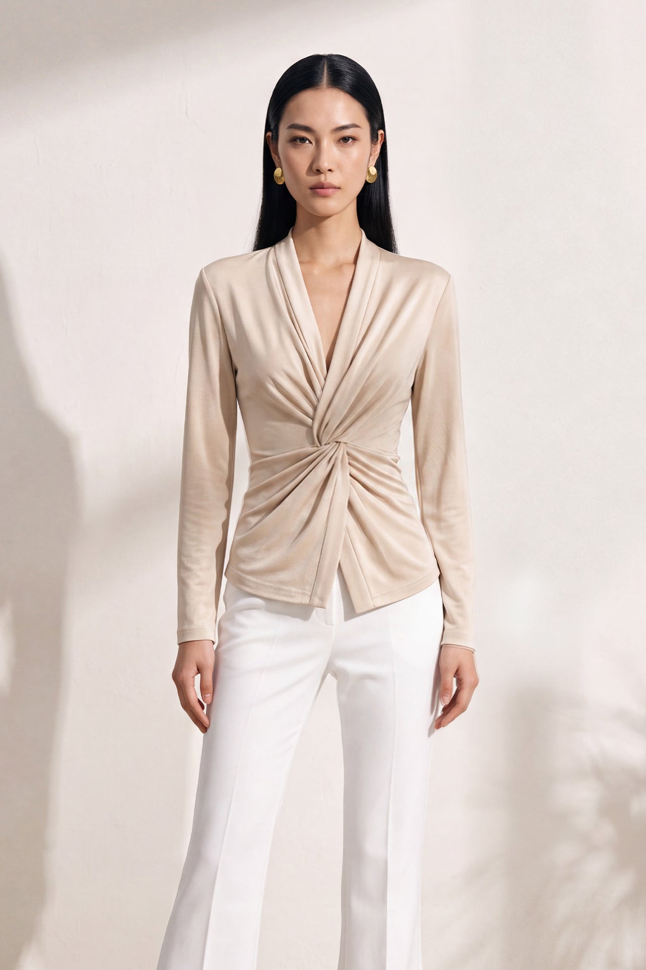 A chic Asian woman modeling an elegant champagne gold long-sleeve top featuring a sophisticated v-neckline and a stylized twist-front knot waist, beautifully paired with high-waisted white trousers.