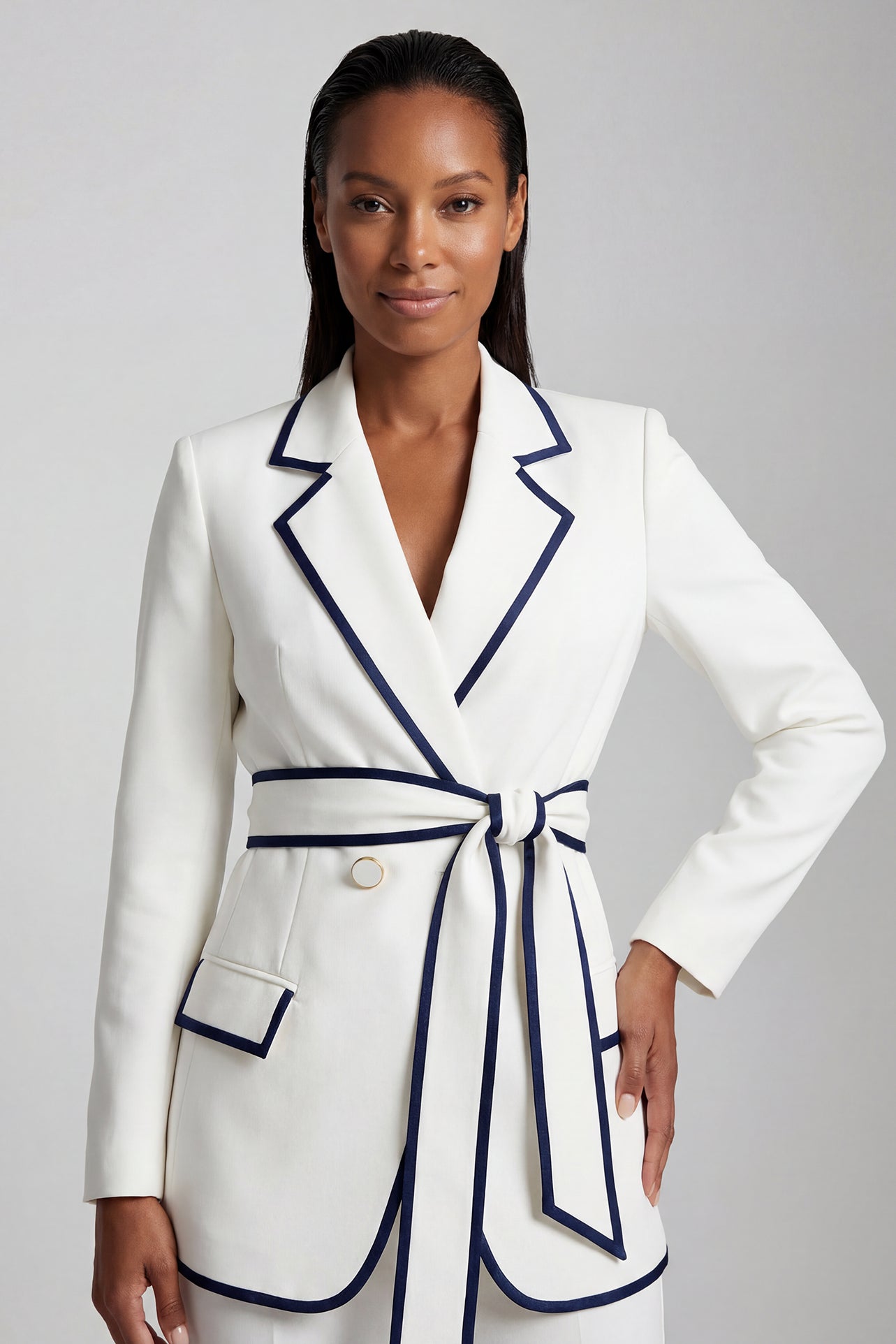 An elegant professional woman wearing a tailored white blazer with navy blue contrast piping, featuring a self-tie belt to define the waist for a smart-casual office look