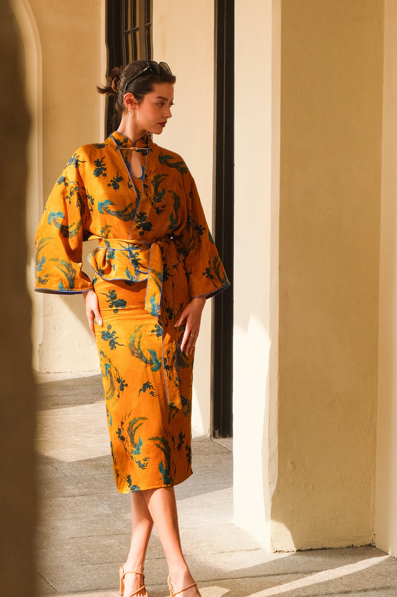 A beautiful woman modeling an elegant mustard yellow modern Chinese jacket, featuring intricate blue phoenix embroidery, a sophisticated mandarin collar, and wide kimono sleeves with a matching waist tie.