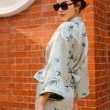 A professional woman modeling an elegant light sage green silk wrap jacket, featuring dark green bamboo leaf prints, a sophisticated mandarin collar with keyhole detail, and wide kimono sleeves.