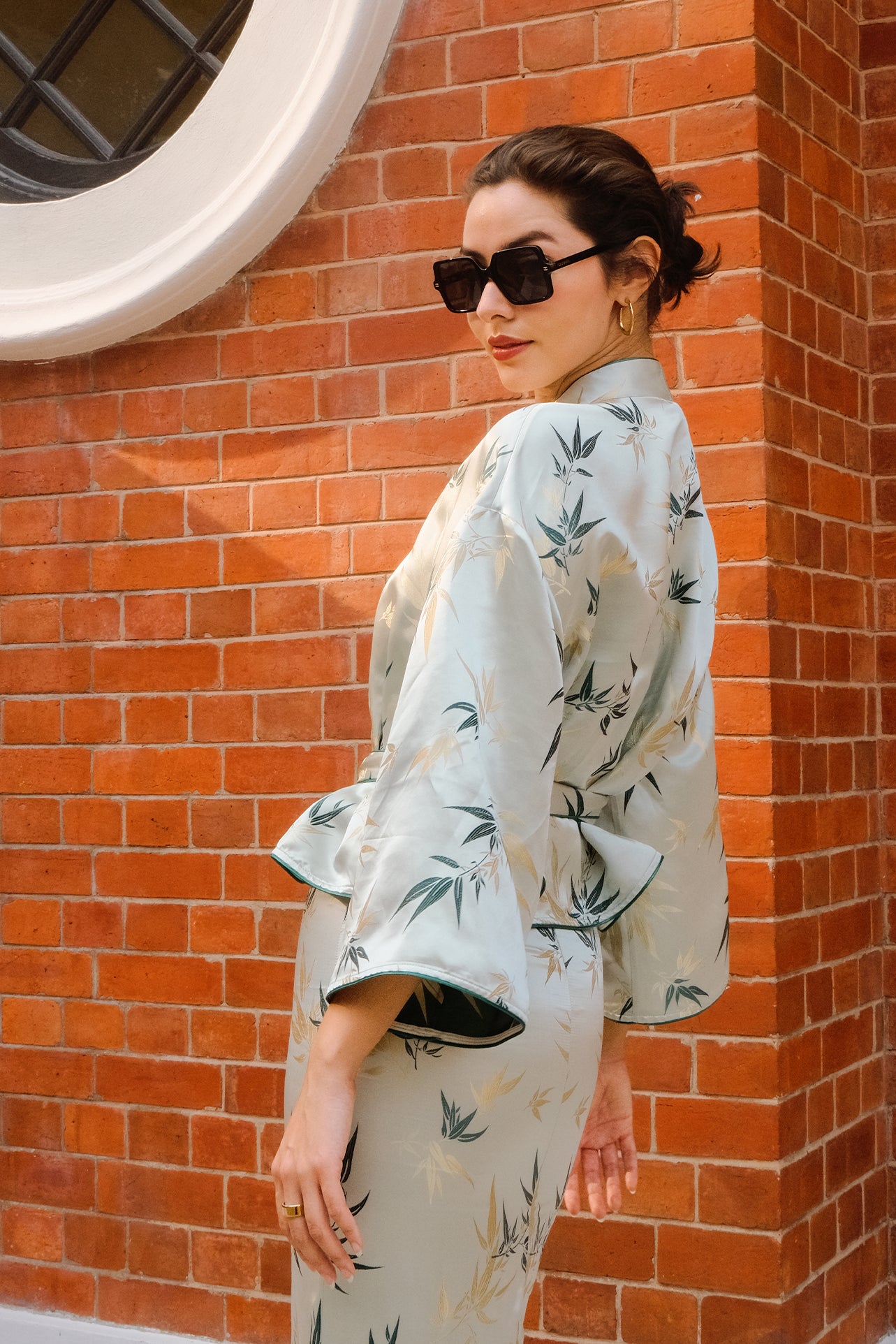 A professional woman modeling an elegant light sage green silk wrap jacket, featuring dark green bamboo leaf prints, a sophisticated mandarin collar with keyhole detail, and wide kimono sleeves.