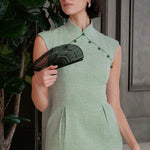 A stylish woman wearing sunglasses and holding a black folding fan, dressed in an elegant mint green sleeveless modern qipao dress featuring a mandarin collar with delicate button trim along the bodice and a tailored midi sheath silhouette.