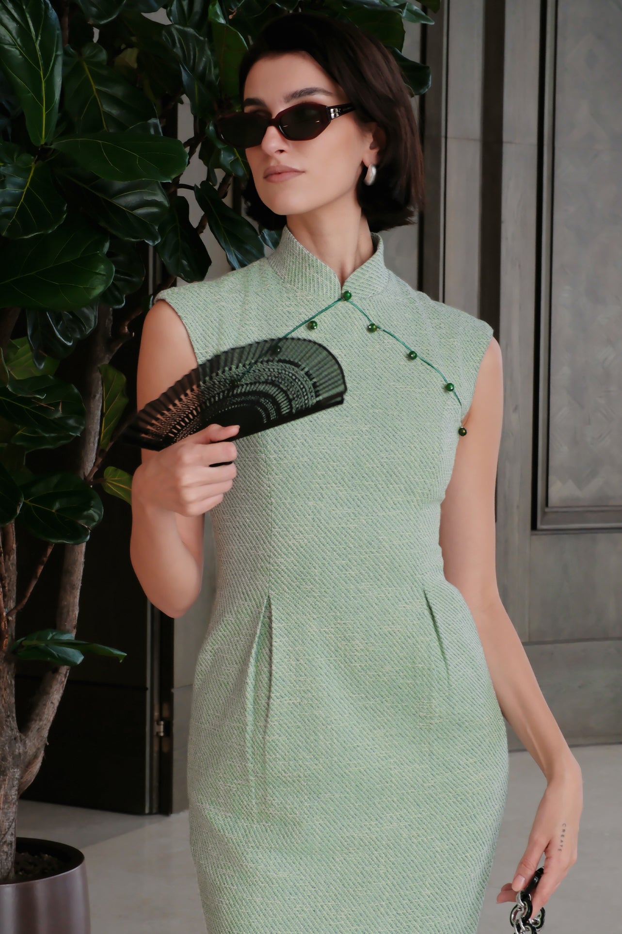 A stylish woman wearing sunglasses and holding a black folding fan, dressed in an elegant mint green sleeveless modern qipao dress featuring a mandarin collar with delicate button trim along the bodice and a tailored midi sheath silhouette.