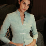 An elegant woman sitting on a modern chair wearing a sophisticated mint green textured suit featuring a cropped jacket with a mandarin collar, three white frog closures, and a matching high-waisted front-slit pencil skirt.