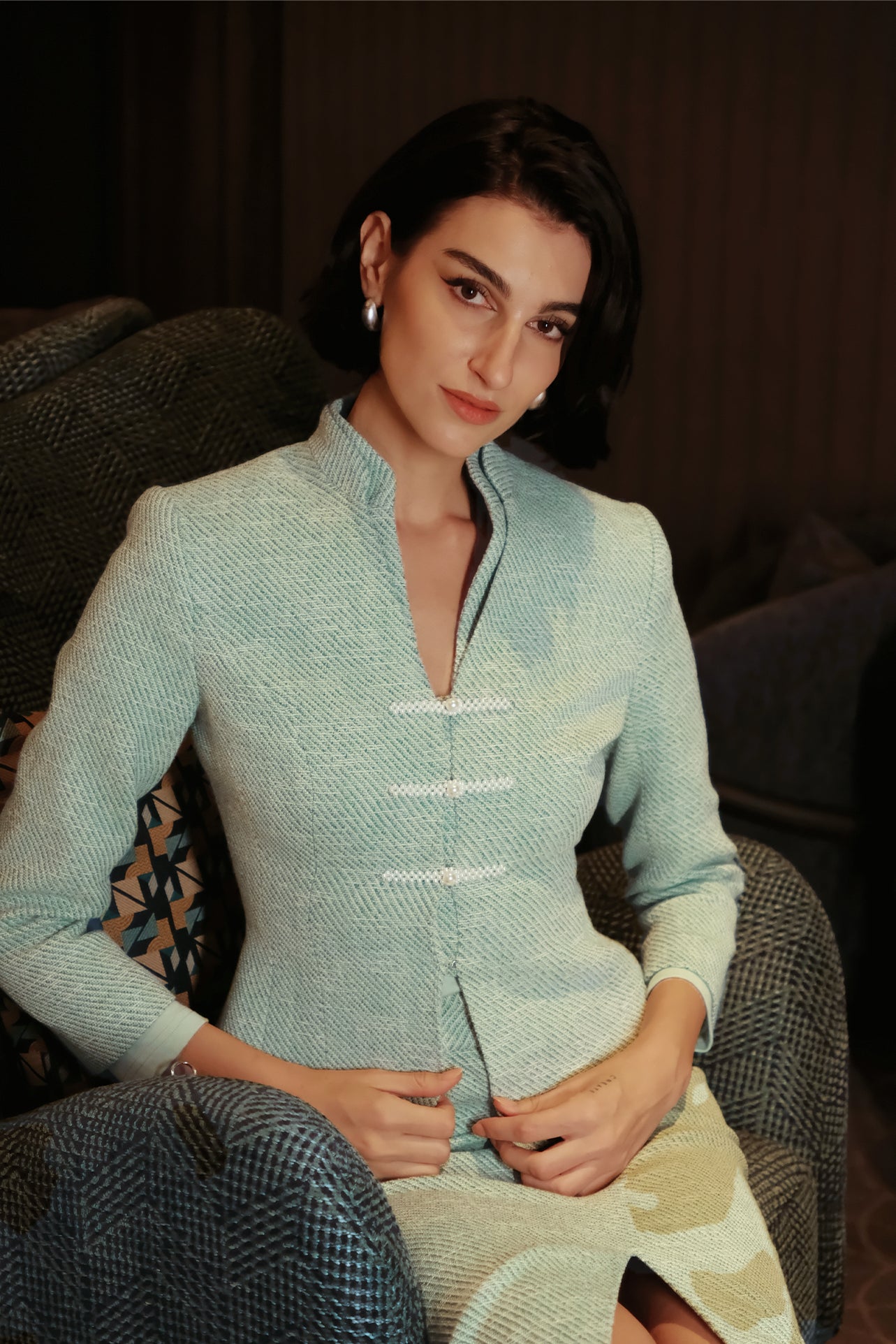 An elegant woman sitting on a modern chair wearing a sophisticated mint green textured suit featuring a cropped jacket with a mandarin collar, three white frog closures, and a matching high-waisted front-slit pencil skirt.