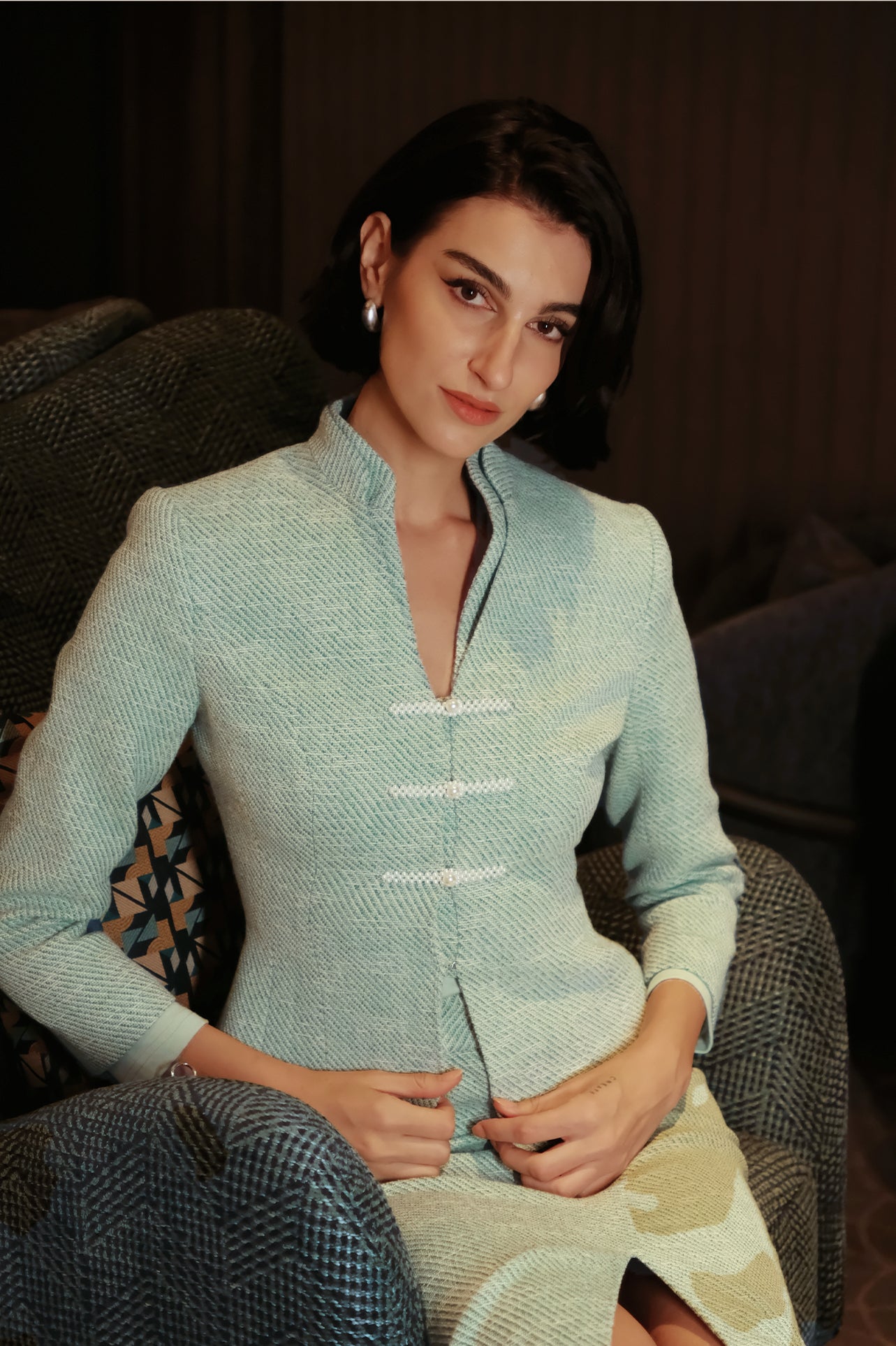 An elegant woman sitting on a modern chair wearing a sophisticated mint green textured suit featuring a cropped jacket with a mandarin collar, three white frog closures, and a matching high-waisted front-slit pencil skirt.
