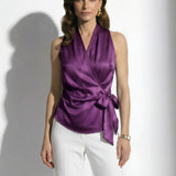 A professional woman modeling elegant satin halter wrap tops in crisp white and deep purple, featuring a sophisticated side bow tie and paired with white high-waisted trousers and a button-front skirt.