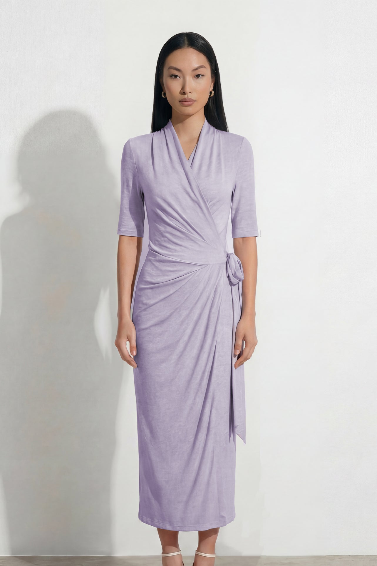 Two professional models wearing elegant wrap midi dresses with elbow-length sleeves; the left in a soft pastel lavender and the right in a deep navy blue, both featuring a sophisticated surplice V-neckline and slimming side ruched draping with a waist tie.