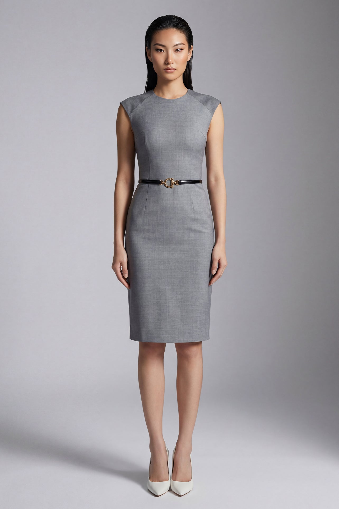 A professional woman wearing a sophisticated grey sleeveless sheath dress, featuring a tailored slim-fit silhouette and an elegant thin black belt with a gold buckle for a polished executive office look.