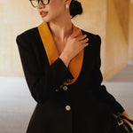 A professional Asian woman wearing glasses and an elegant black tailored blazer featuring a striking mustard yellow contrast shawl lapel, statement front button, and buttoned cuffs for a sophisticated intellectual corporate look.