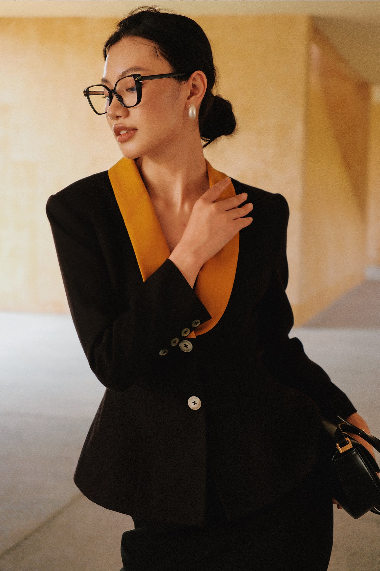 A professional Asian woman wearing glasses and an elegant black tailored blazer featuring a striking mustard yellow contrast shawl lapel, statement front button, and buttoned cuffs for a sophisticated intellectual corporate look.