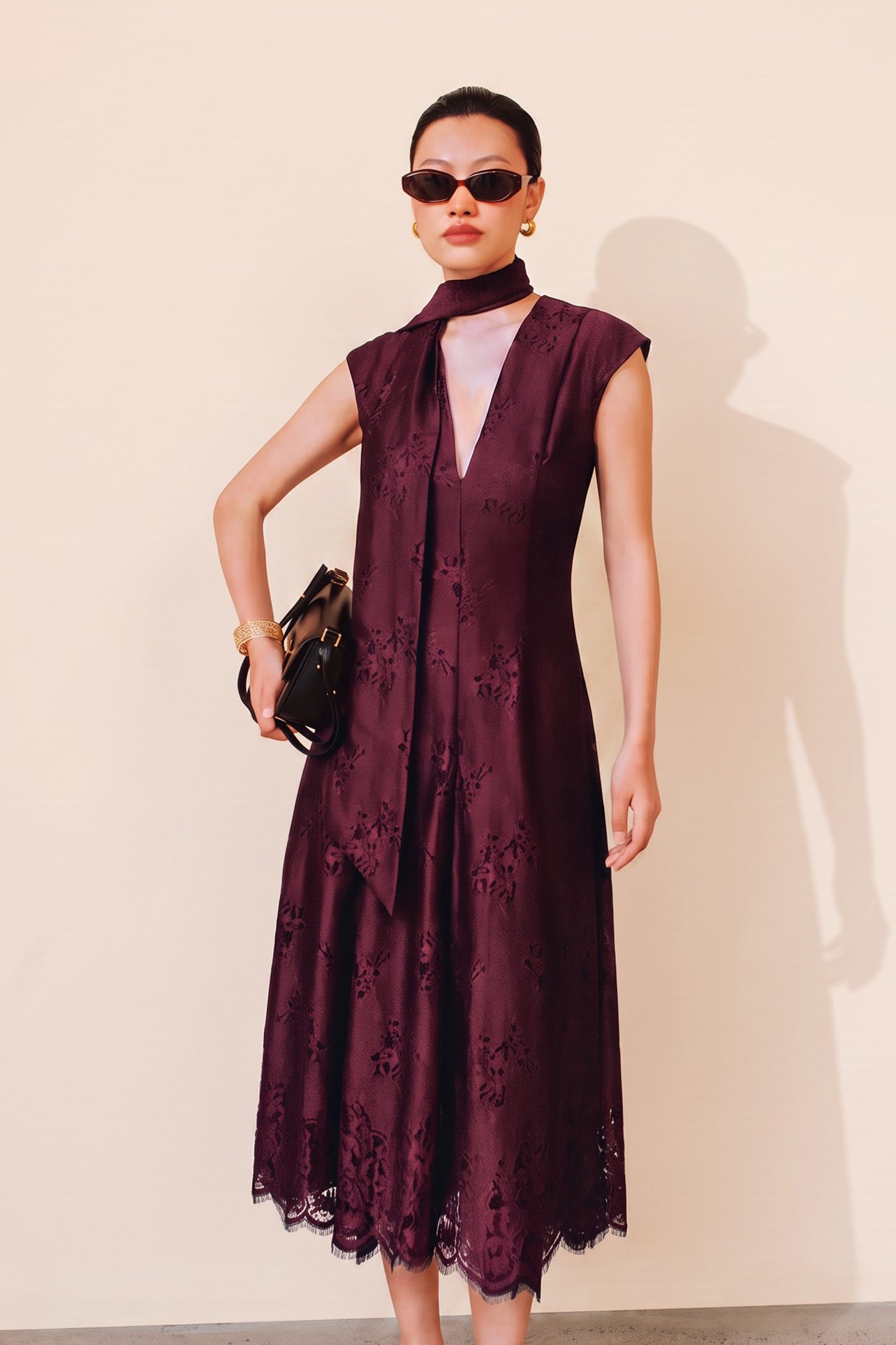 A chic woman wearing sunglasses and an elegant deep burgundy sleeveless V-neck A-line midi dress featuring textured jacquard fabric, delicate lace hemline, and a matching flowing neck scarf.