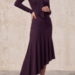 An elegant woman wearing a sophisticated deep plum-purple long-sleeve knit midi dress featuring a draped high-neck tie scarf and a dramatic asymmetrical flounce mermaid hemline.