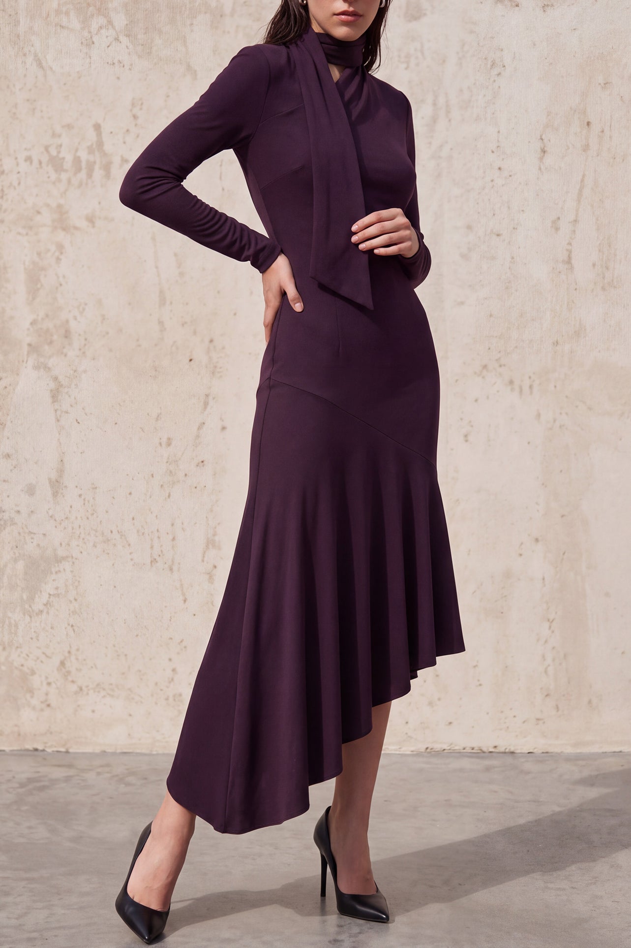 An elegant woman wearing a sophisticated deep plum-purple long-sleeve knit midi dress featuring a draped high-neck tie scarf and a dramatic asymmetrical flounce mermaid hemline.