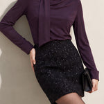 A chic woman wearing an elegant deep plum long-sleeve blouse with an asymmetric high-neck scarf tie, neatly tucked into a high-waisted black tweed skirt embellished with subtle sparkling sequins.