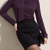A chic woman wearing an elegant deep plum long-sleeve blouse with an asymmetric high-neck scarf tie, neatly tucked into a high-waisted black tweed skirt embellished with subtle sparkling sequins.