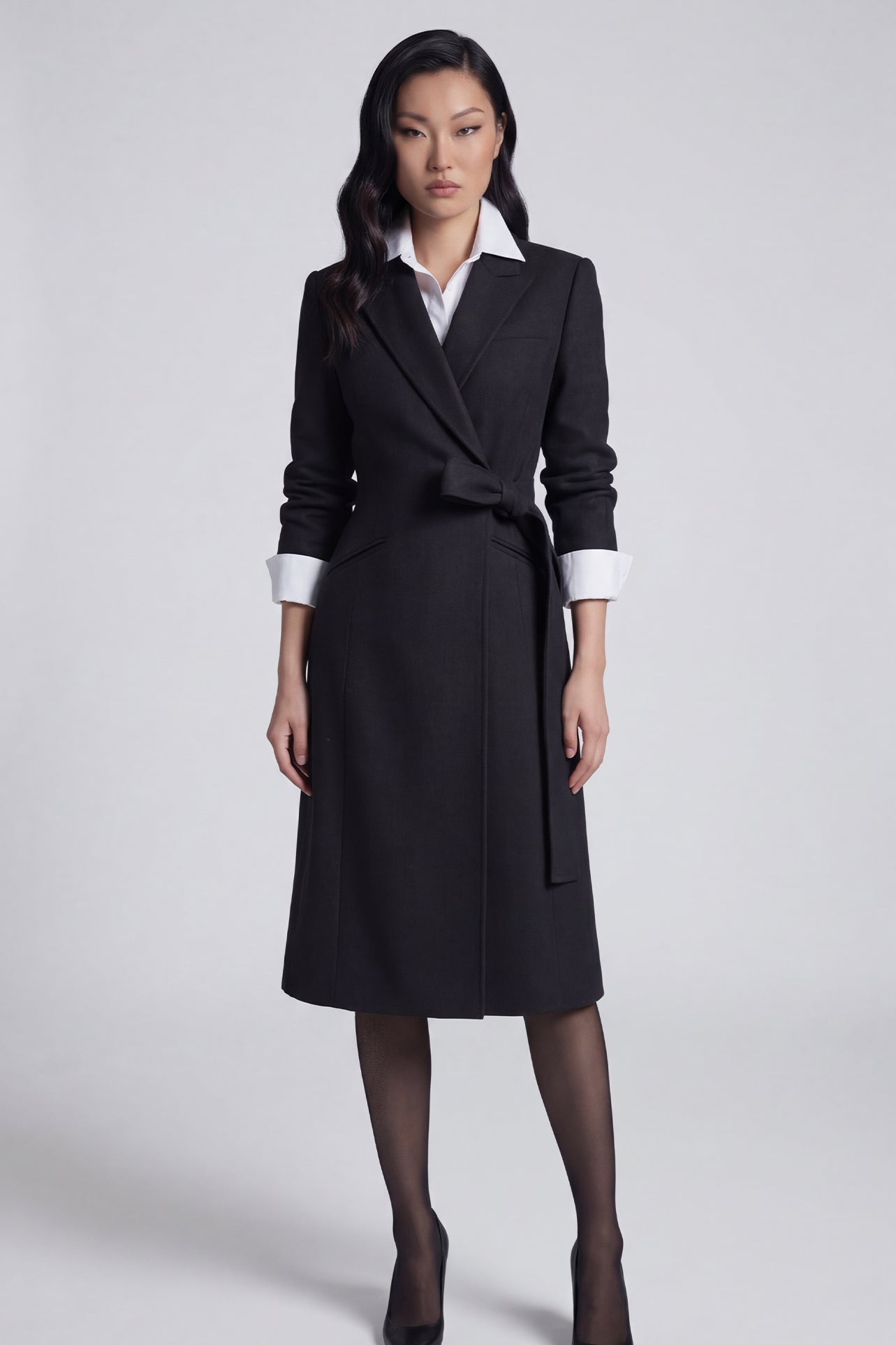A professional woman wearing a sophisticated black long-sleeve coat dress, featuring an elegant tie-waist belt and notched lapels, styled over a white shirt for a polished executive business look.
