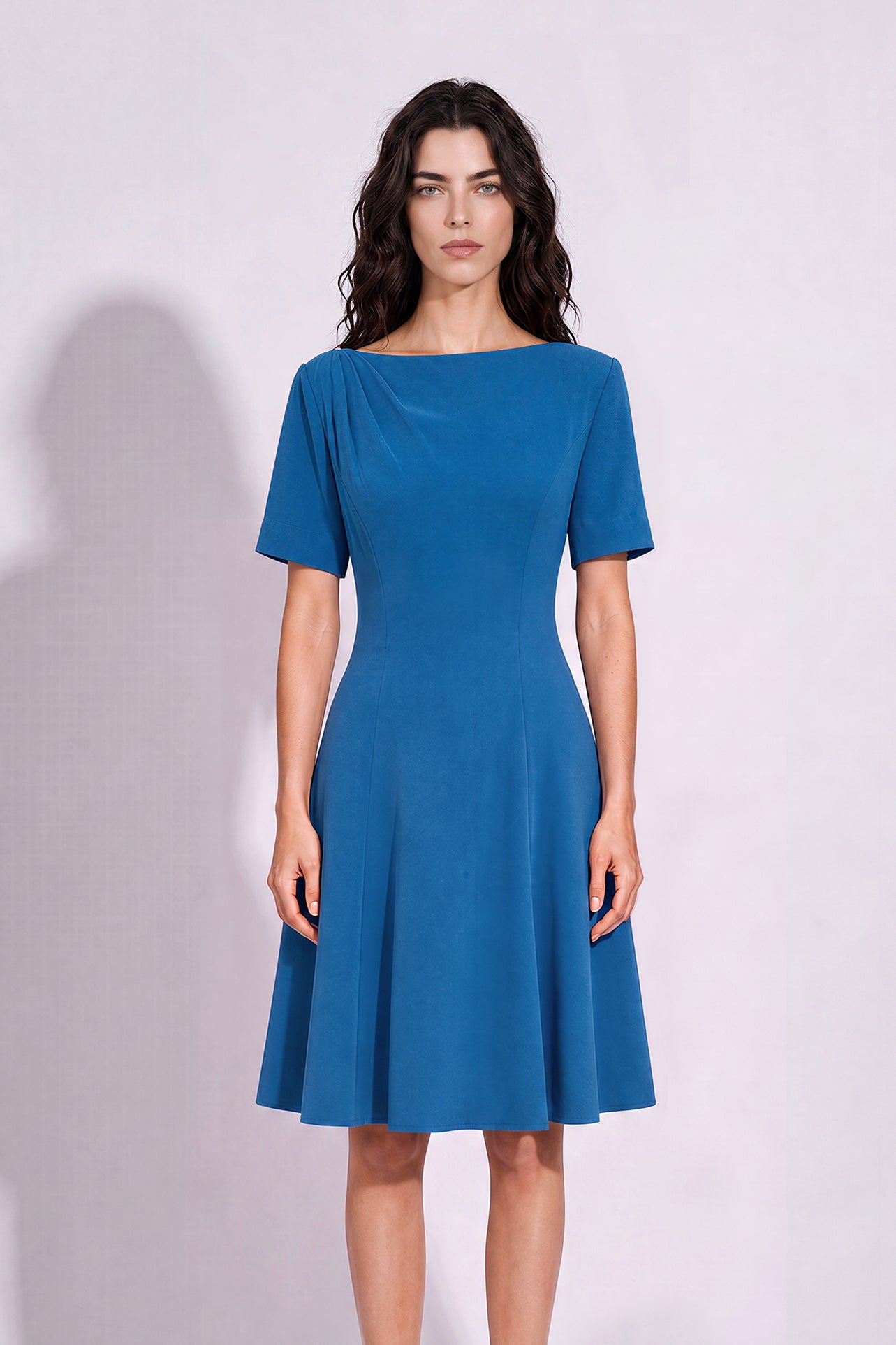 A beautiful model wearing a sophisticated medium blue A-line midi dress featuring an elegant boat neckline with refined asymmetric shoulder pleating, short sleeves, and a flowing flared swing skirt.