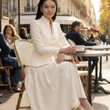 A sophisticated woman enjoying coffee in Paris wearing an elegant cream white modern qipao suit, featuring a mandarin collar blazer with traditional frog buttons and a matching pleated skirt for a quiet luxury oriental look.
