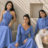 A model showcasing an elegant blue and white vertical striped maxi dress, featuring a sophisticated criss-cross halter neckline, a structured wide banded waist for a slimming silhouette, and a long flowing skirt, paired with white strap high heels.