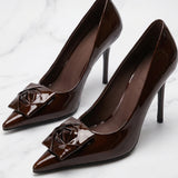 A pair of elegant dark chocolate brown patent leather pumps featuring a pointed toe and a sophisticated 3D rose floral decoration on the front, perfect for a quiet luxury office or evening look.