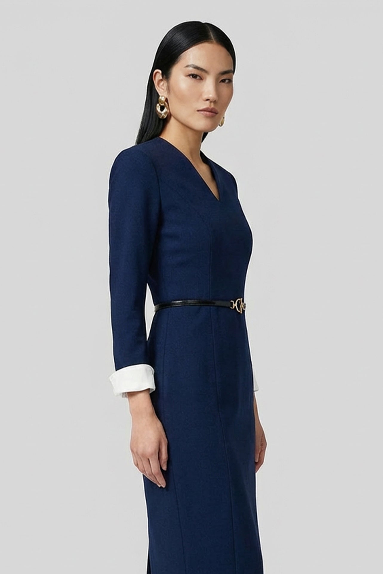 A professional woman wearing a sophisticated navy blue V-neck sheath dress, featuring elegant white contrast turn-up cuffs and a slim black belt with a gold buckle for a polished executive look.