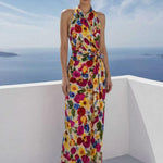A beautiful professional model presenting an elegant tropical floral print halter maxi dress with a blue ocean background; featuring a sophisticated wrap tie-waist silhouette and vibrant multicolored botanical patterns for a chic summer resort look.