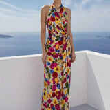 A beautiful professional model presenting an elegant tropical floral print halter maxi dress with a blue ocean background; featuring a sophisticated wrap tie-waist silhouette and vibrant multicolored botanical patterns for a chic summer resort look.