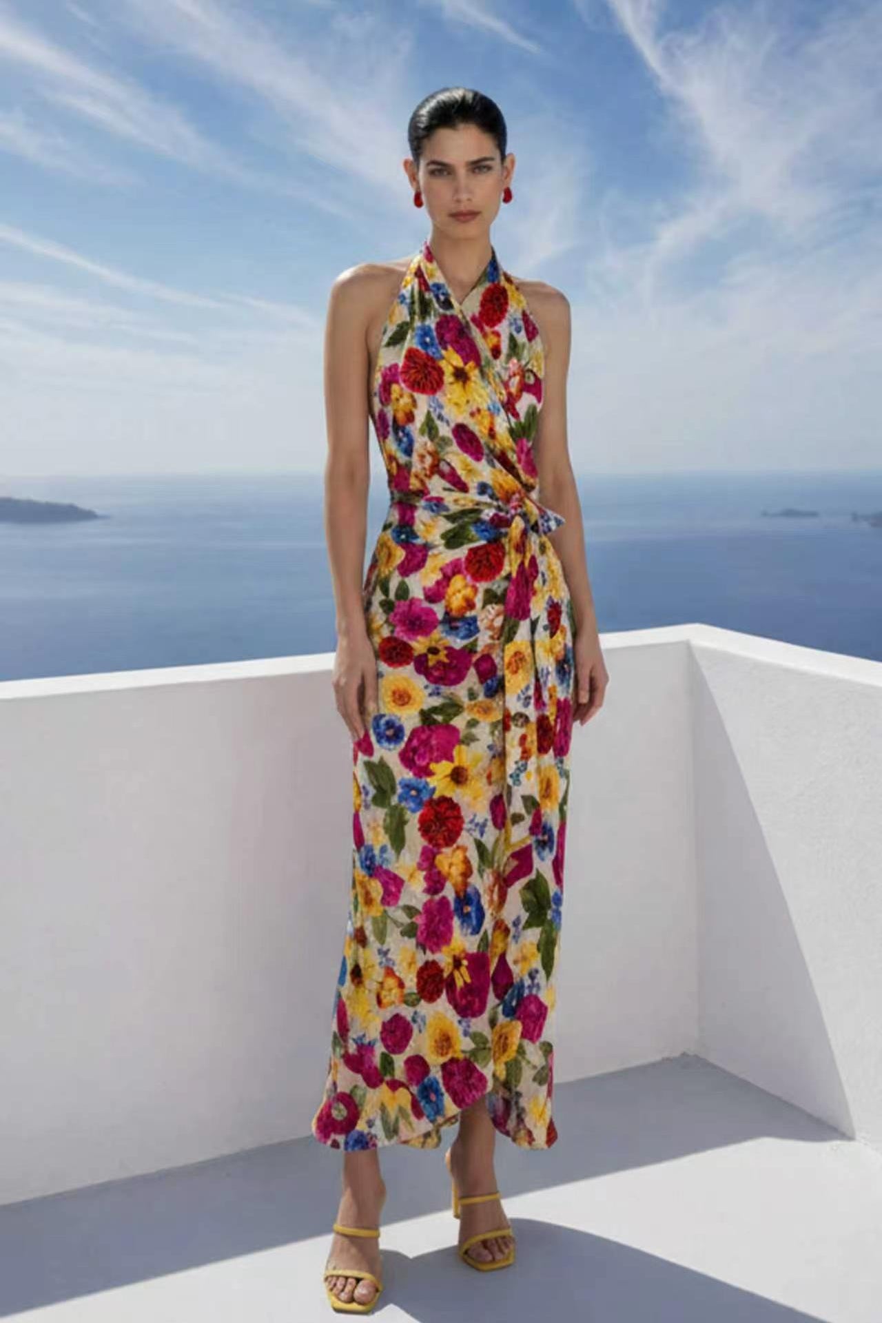 A beautiful professional model presenting an elegant tropical floral print halter maxi dress with a blue ocean background; featuring a sophisticated wrap tie-waist silhouette and vibrant multicolored botanical patterns for a chic summer resort look.