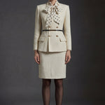 A professional woman wearing a sophisticated cream white tweed skirt suit, featuring a double-breasted blazer with gold buttons, a slim black belt, and an elegant polka dot pussy bow tie for a polished French chic office look.