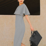 A stylish woman walking on a city street wearing an elegant light grey sheath midi dress with a sophisticated capelet sleeve design, round neckline, and side slit, carrying a black leather handbag.