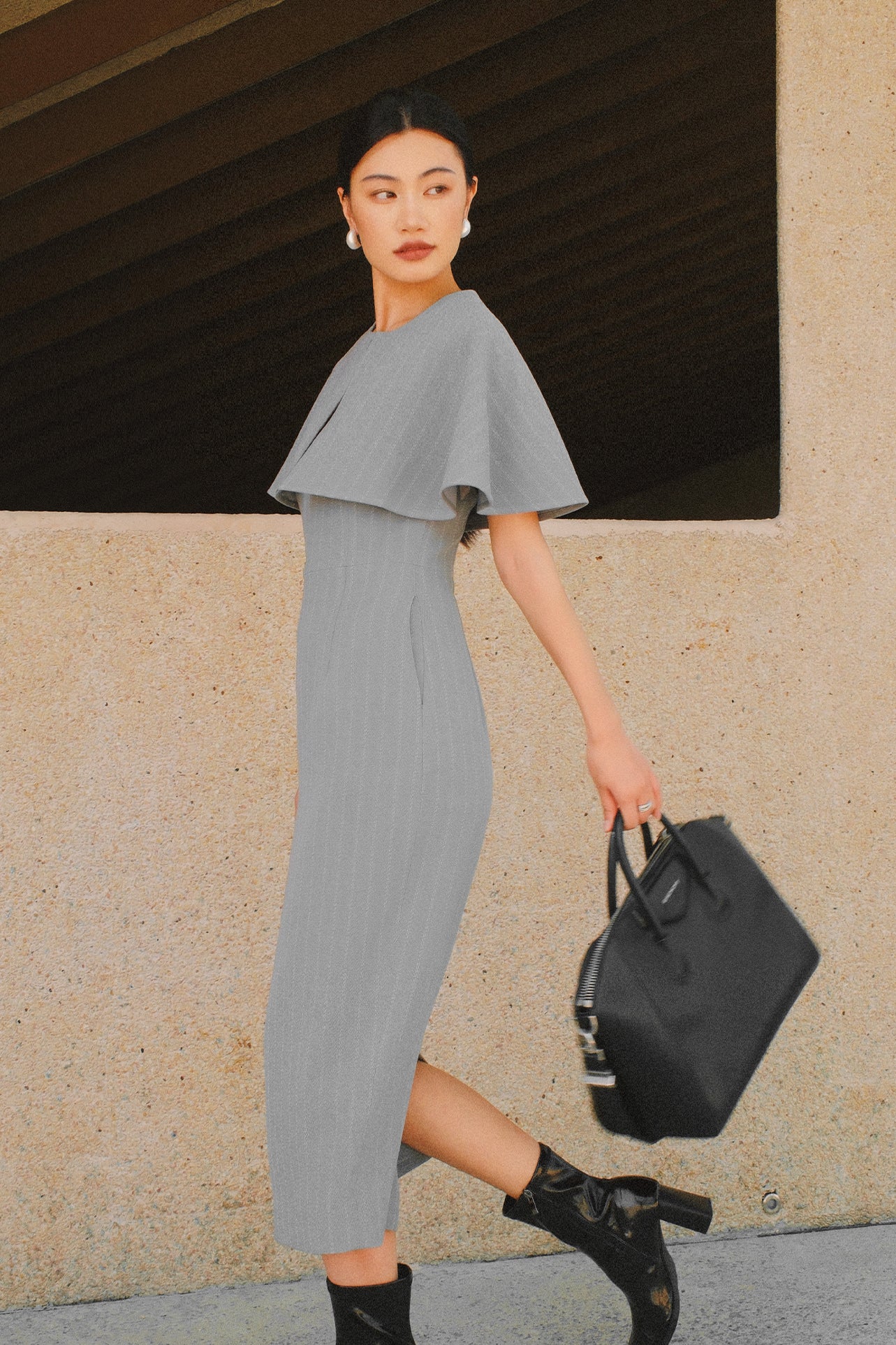 A stylish woman walking on a city street wearing an elegant light grey sheath midi dress with a sophisticated capelet sleeve design, round neckline, and side slit, carrying a black leather handbag.