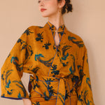 A beautiful woman modeling an elegant mustard yellow modern Chinese jacket, featuring intricate blue phoenix embroidery, a sophisticated mandarin collar, and wide kimono sleeves with a matching waist tie.