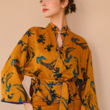 A beautiful woman modeling an elegant mustard yellow modern Chinese jacket, featuring intricate blue phoenix embroidery, a sophisticated mandarin collar, and wide kimono sleeves with a matching waist tie.