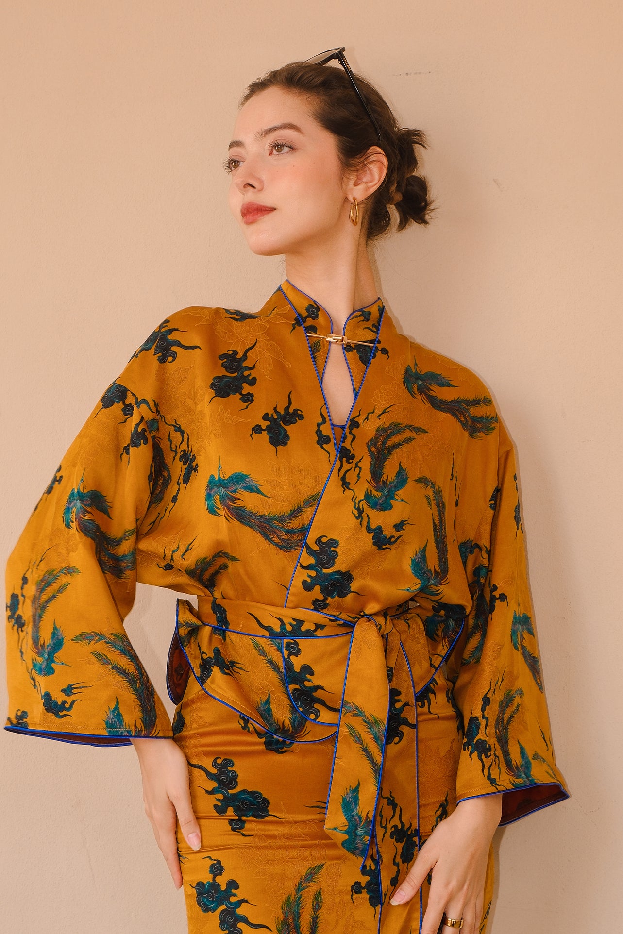 A beautiful woman modeling an elegant mustard yellow modern Chinese jacket, featuring intricate blue phoenix embroidery, a sophisticated mandarin collar, and wide kimono sleeves with a matching waist tie.