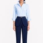 A model showcasing elegant navy blue high-waisted work trousers, featuring a unique curved overlay panel design at the waist, a slim golden metal bar buckle, pressed front creases, and chic cuffed hems, paired with a light blue button-up shirt.