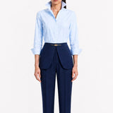 A model showcasing elegant navy blue high-waisted work trousers, featuring a unique curved overlay panel design at the waist, a slim golden metal bar buckle, pressed front creases, and chic cuffed hems, paired with a light blue button-up shirt.