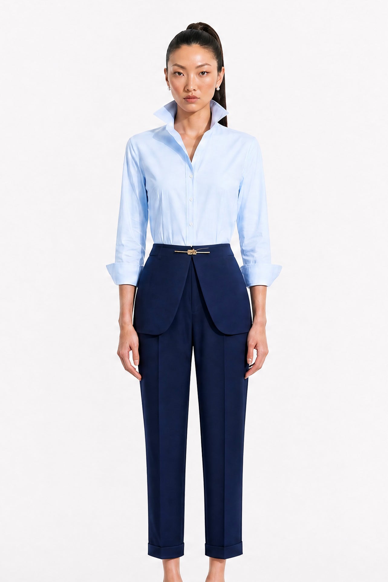 A model showcasing elegant navy blue high-waisted work trousers, featuring a unique curved overlay panel design at the waist, a slim golden metal bar buckle, pressed front creases, and chic cuffed hems, paired with a light blue button-up shirt.