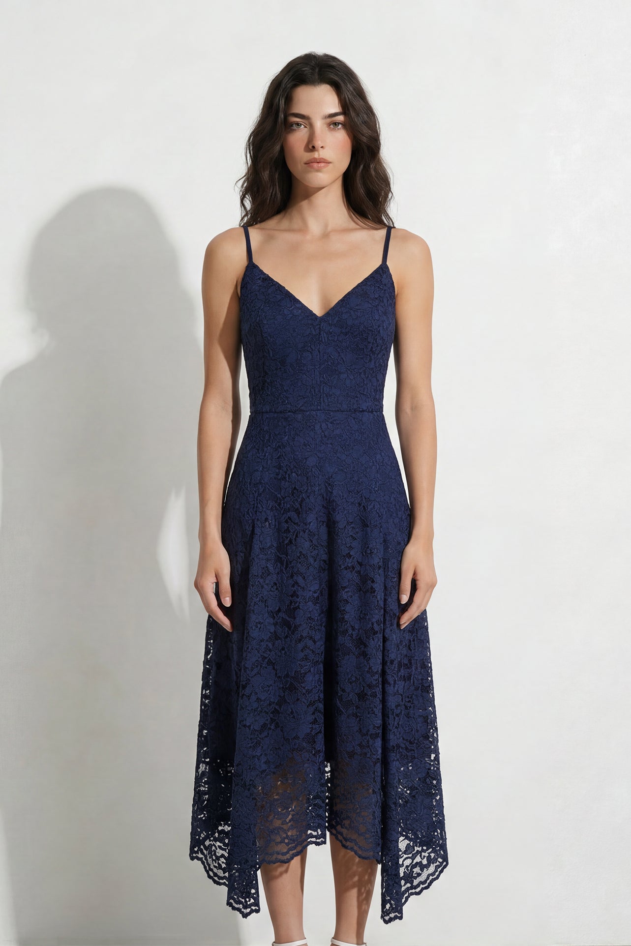 A professional woman modeling an elegant navy blue lace midi dress, featuring delicate V-neck spaghetti straps, a tailored fit-and-flare silhouette, and a beautiful flowing asymmetric handkerchief lace trim hem.