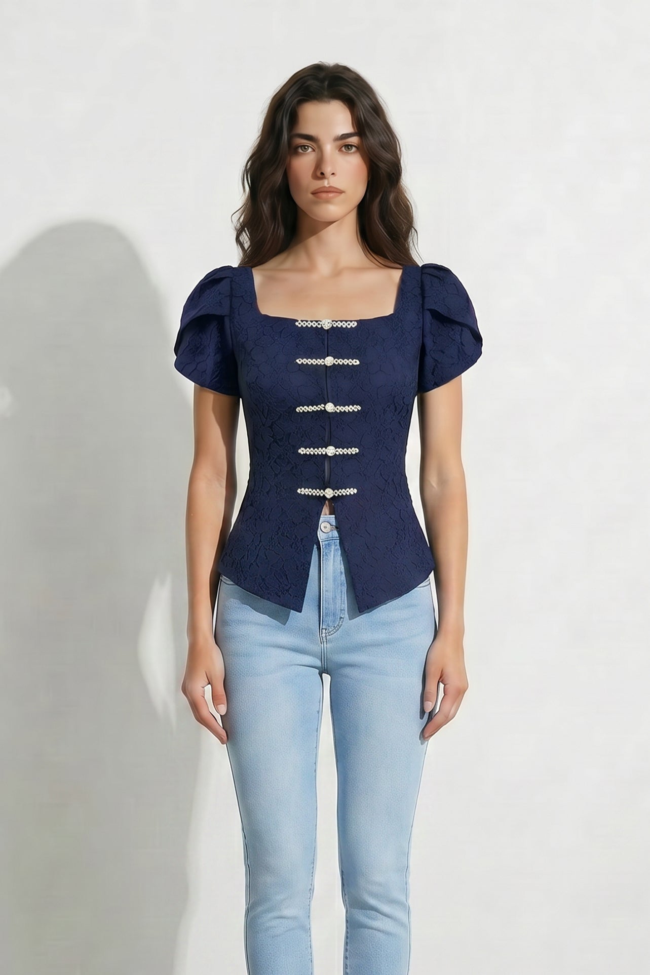 A professional woman modeling an elegant navy blue lace short top, featuring a retro square neckline, romantic short puff sleeves, and a series of pearl and rhinestone frog closures on the front with a tailored triangular split hem, paired with light blue high-waisted jeans.