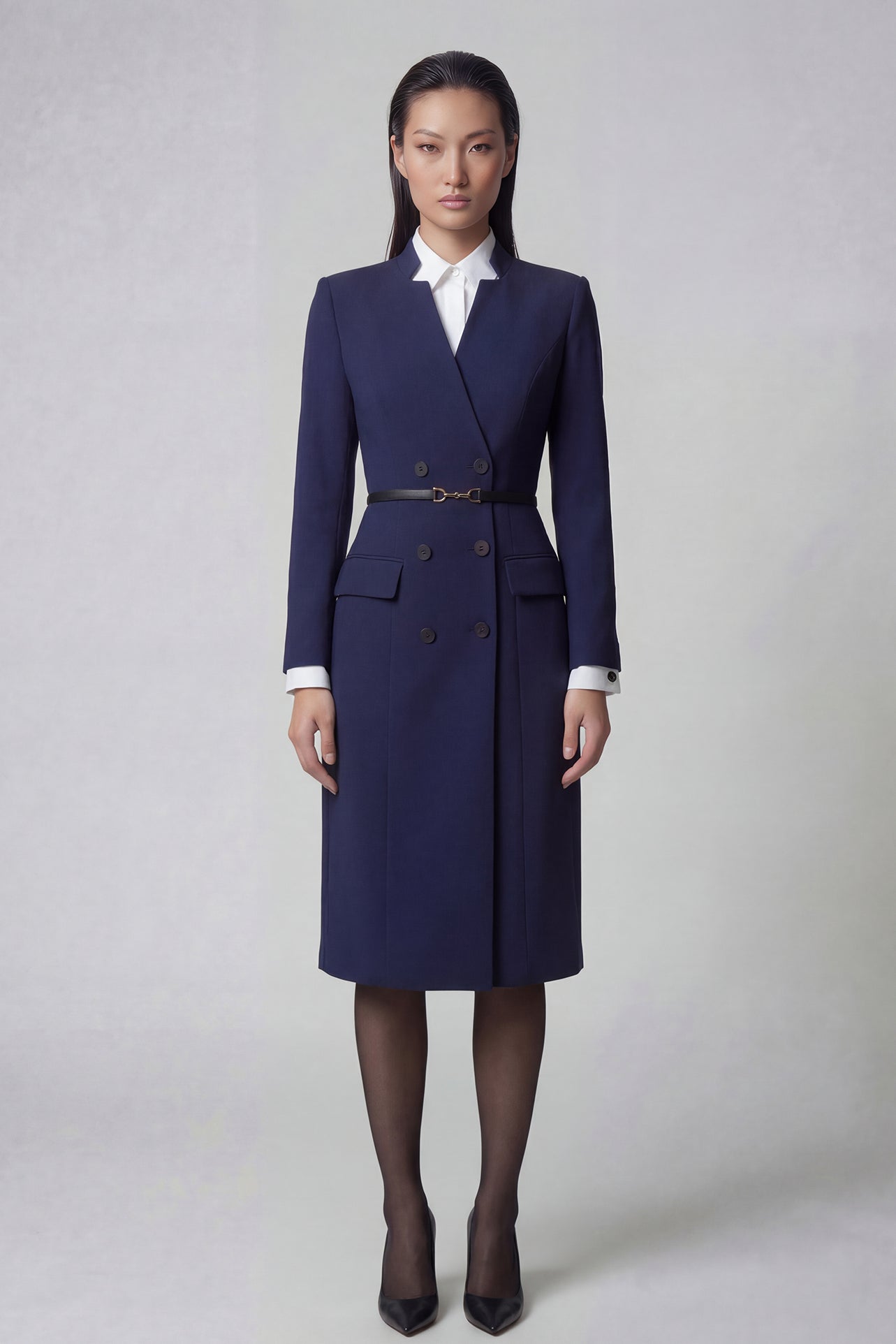 An elegant professional woman wearing a sophisticated navy blue double-breasted long coat dress, featuring a black waist belt and structured shoulders for a powerful executive look.