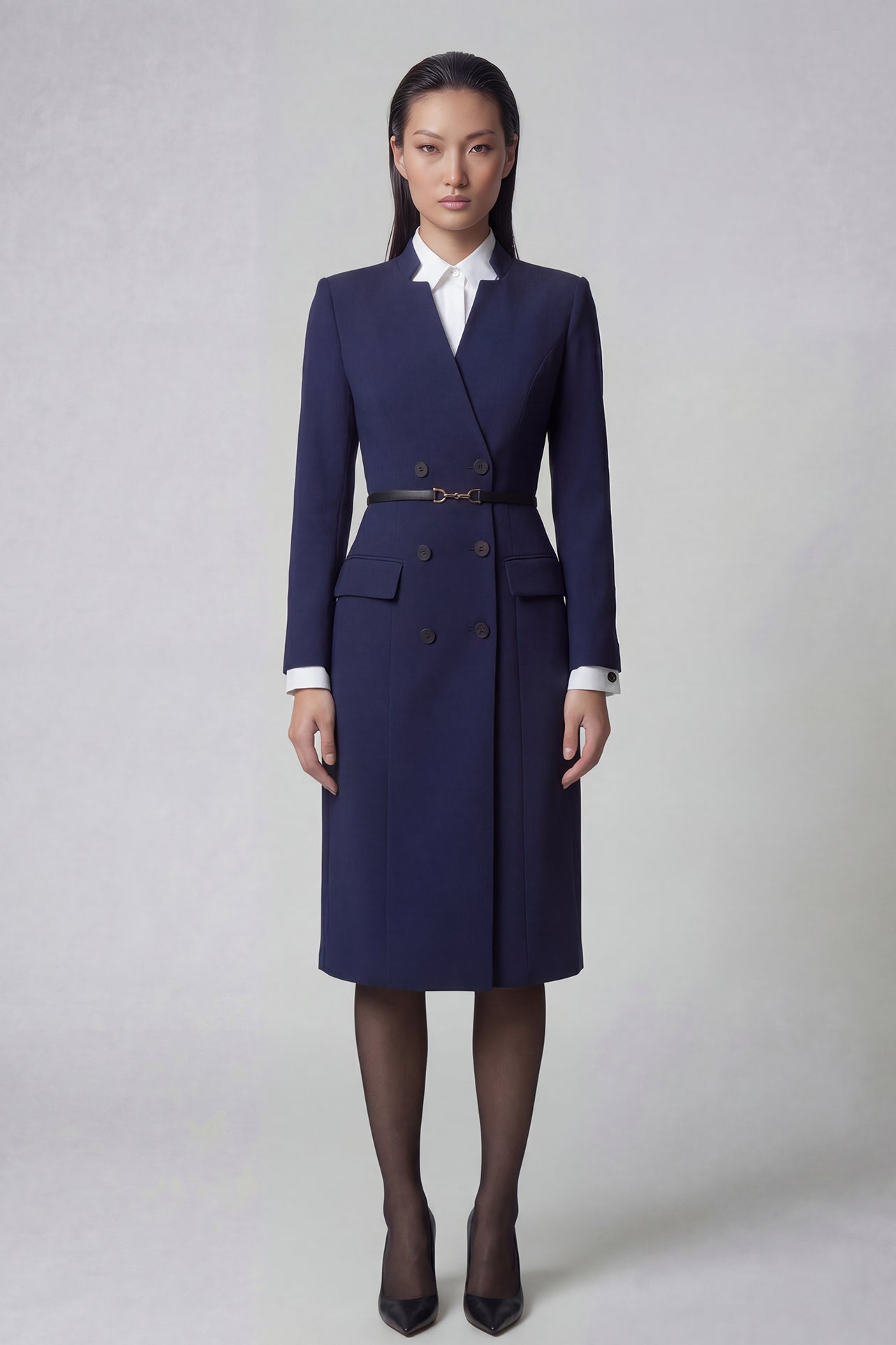 An elegant professional woman wearing a sophisticated navy blue double-breasted long coat dress, featuring a black waist belt and structured shoulders for a powerful executive look.