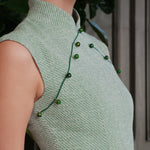 A stylish woman wearing sunglasses and holding a black folding fan, dressed in an elegant mint green sleeveless modern qipao dress featuring a mandarin collar with delicate button trim along the bodice and a tailored midi sheath silhouette.