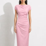 A professional woman modeling an elegant pastel pink satin midi dress, featuring a sophisticated cowl neckline, delicate cap sleeves, and slimming asymmetric ruched drapes across the waist for a quiet luxury look.