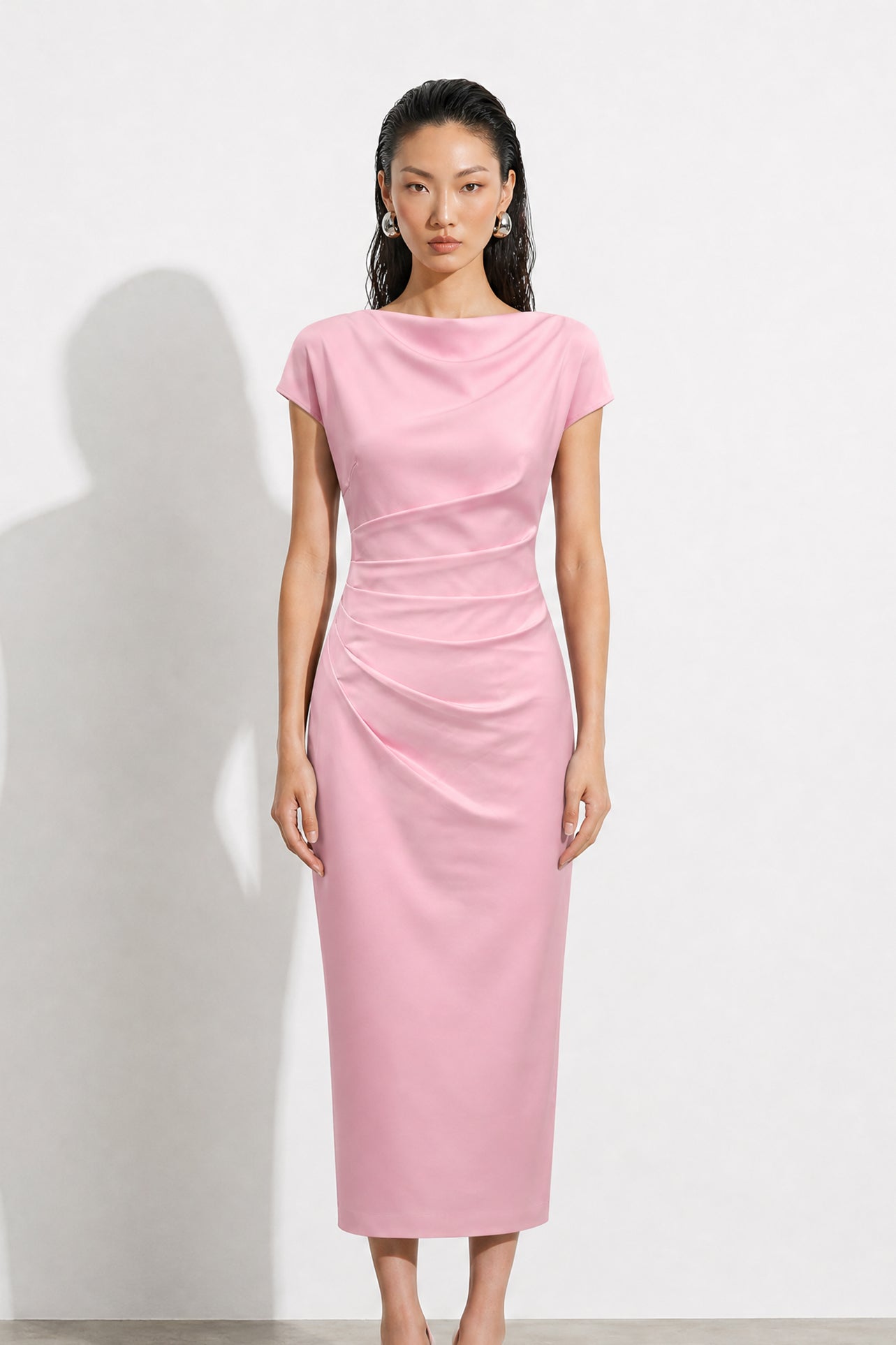 A professional woman modeling an elegant pastel pink satin midi dress, featuring a sophisticated cowl neckline, delicate cap sleeves, and slimming asymmetric ruched drapes across the waist for a quiet luxury look.
