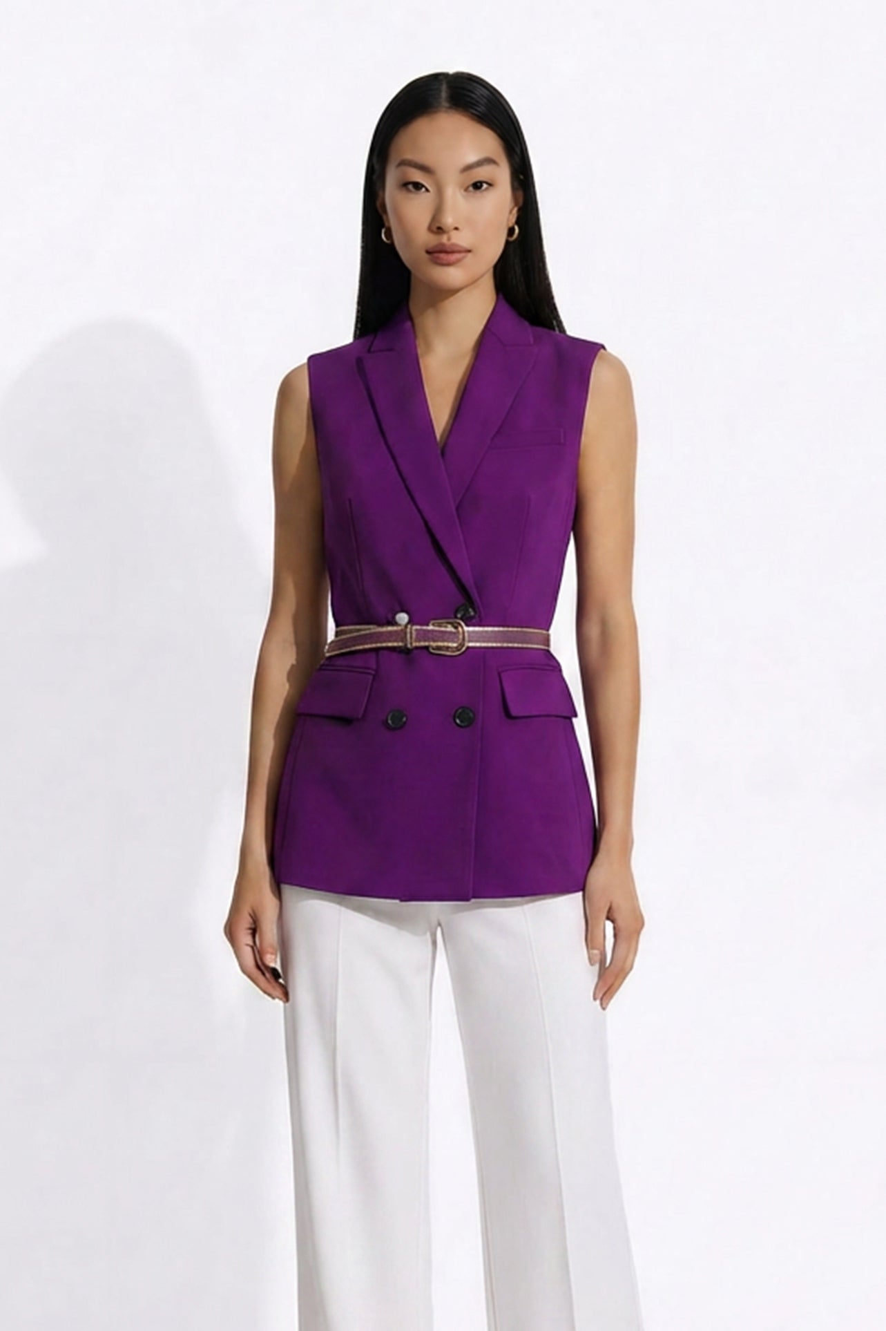 A professional woman modeling an elegant deep purple sleeveless double-breasted blazer vest, featuring a notched lapel, front flap pockets, and a slim belt, paired with white tailored trousers for a sophisticated office look.