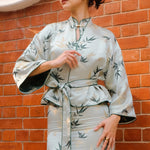 A professional woman modeling an elegant light sage green silk wrap jacket, featuring dark green bamboo leaf prints, a sophisticated mandarin collar with keyhole detail, and wide kimono sleeves.