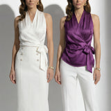 A professional woman modeling elegant satin halter wrap tops in crisp white and deep purple, featuring a sophisticated side bow tie and paired with white high-waisted trousers and a button-front skirt.